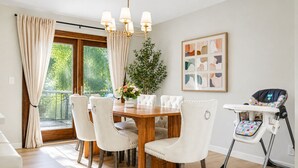 A dining area with a wooden table, white upholstered chairs, a baby high chair, a chandelier, a potted plant, and a window with curtains. - The Poolside farm house (Omaha)