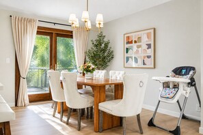 Ein Essbereich mit Holztisch, weiß gepolsterten Stühlen, einem Hochstuhl, einer Pendelleuchte, einem Topfpflanzen und einem fenster mit Vorhängen.