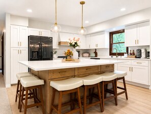 A modern kitchen with white cabinetry, a black refrigerator, a large island with stools, pendant lighting, and a window with a view.