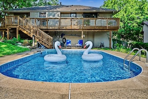 A swimming pool with two swan-shaped inflatable floats, a wooden deck, and a two-story house with a covered patio.