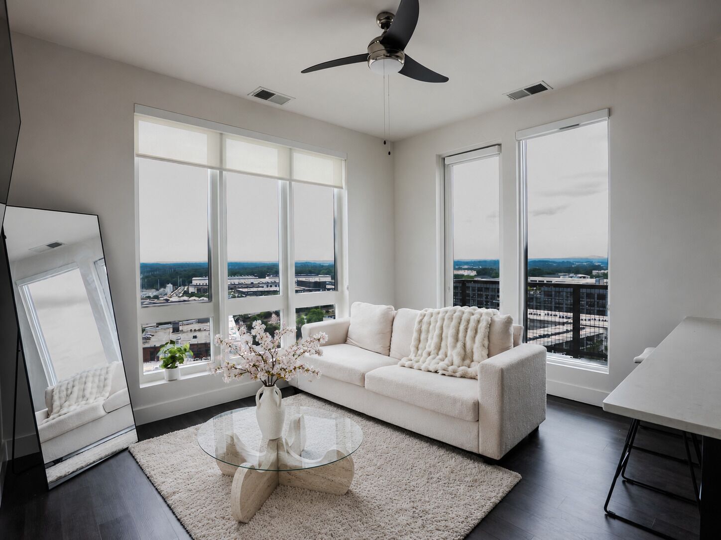 A modern living room with a white sofa, a glass coffee table, and a view of a cityscape through large windows.
