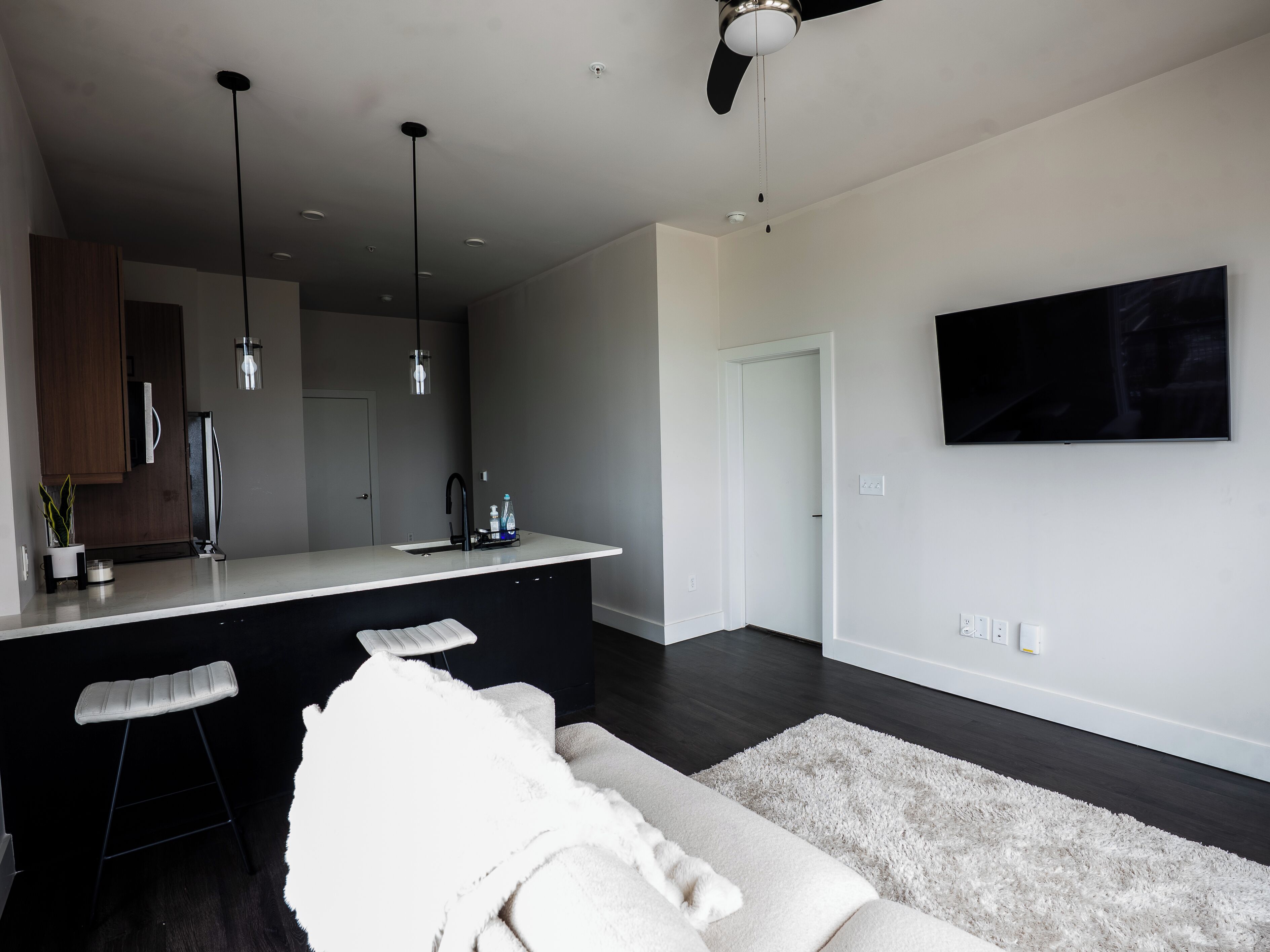 A modern kitchen with a central island, pendant lights, and a large flat-screen TV mounted on the wall.