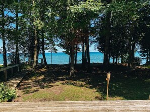 A wooden deck leading to a grassy area with trees and a view of the water. - Lake Huron Cottage| Beach, Firepit + Sauna Retreat (Cheboygan)
