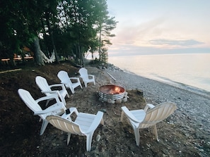 A beachside seating area with white chairs arranged around a fire pit. - Lake Huron Cottage| Beach, Firepit + Sauna Retreat (Cheboygan)