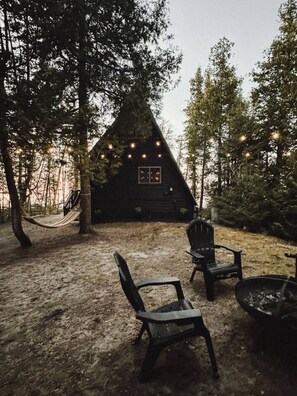 A cabin with a triangular roof surrounded by trees, a hammock, and outdoor seating.