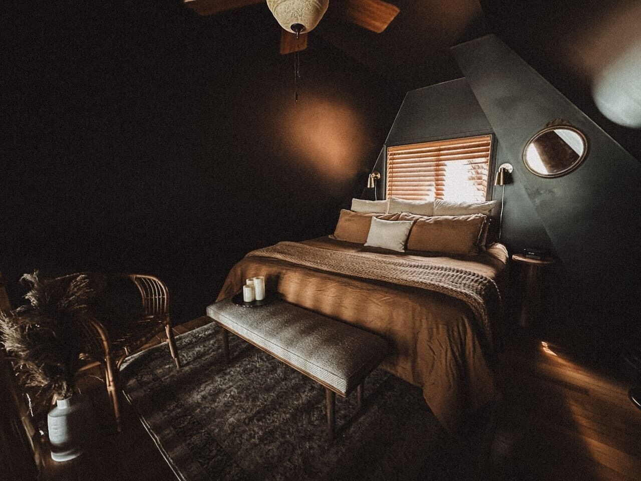 A dimly lit bedroom with a bed, a bench, a chair, and a round mirror on the wall.
