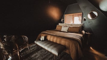 A dimly lit bedroom with a bed, a bench, a chair, and a round mirror on the wall.