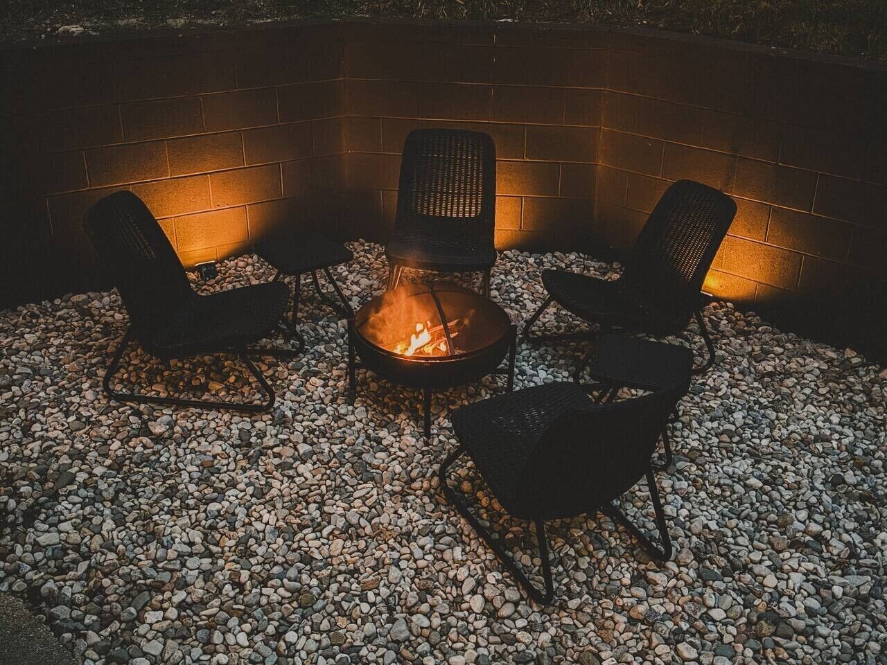 A fire pit with three chairs arranged around it, set on a gravel surface with a brick wall in the background.