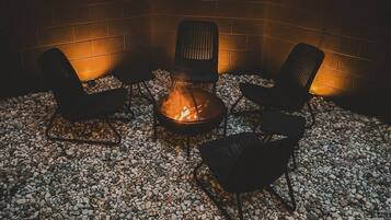 A fire pit with three chairs arranged around it, set on a gravel surface with a brick wall in the background.