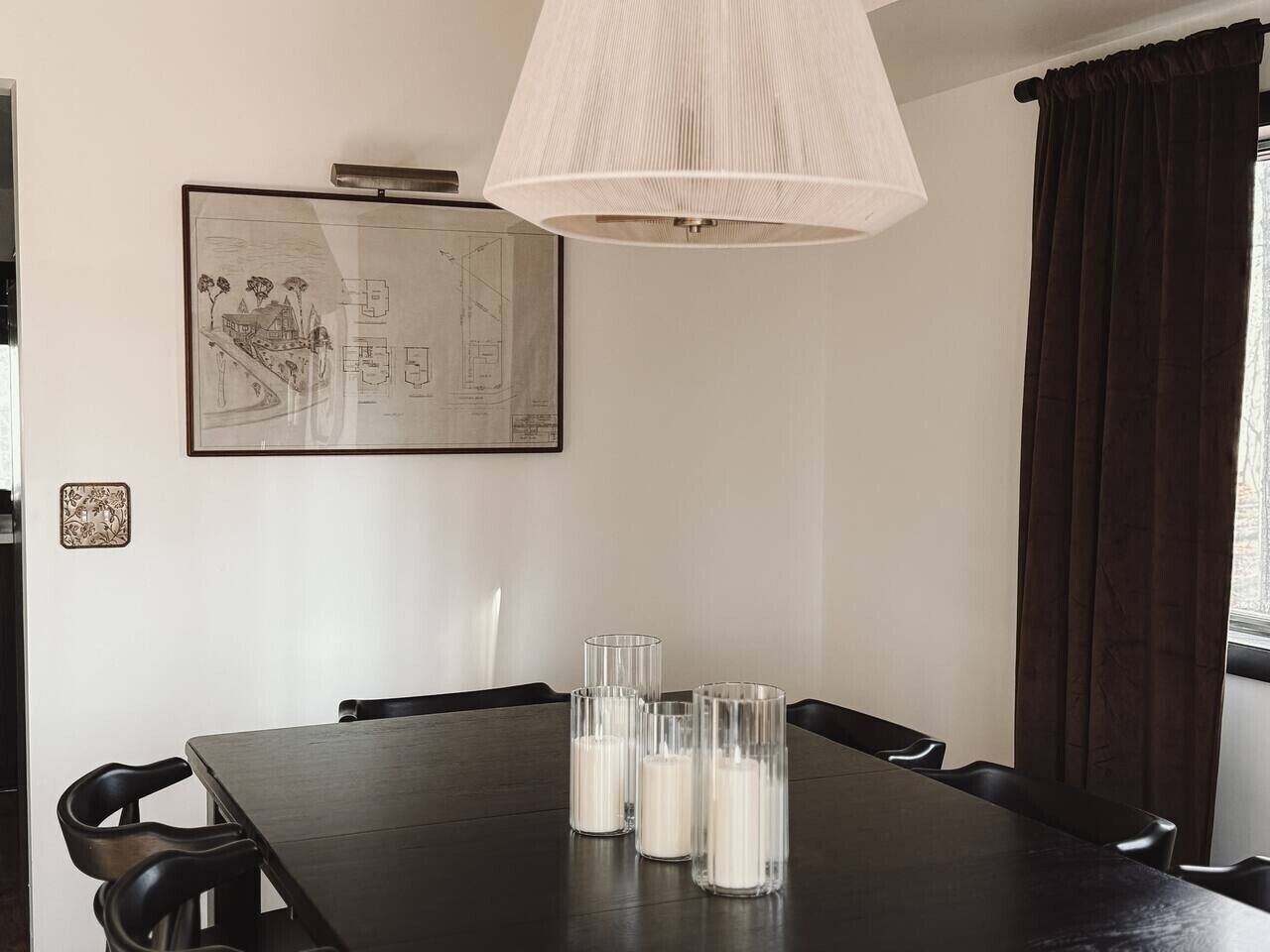 Un comedor con una mesa de madera oscura, cuatro vasos transparentes con velas, una lámpara colgante y un plano arquitectónico enmarcado en la pared.
