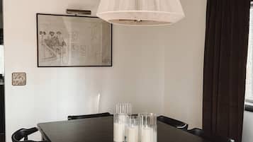 Un comedor con una mesa de madera oscura, cuatro vasos transparentes con velas, una lámpara colgante y un plano arquitectónico enmarcado en la pared.