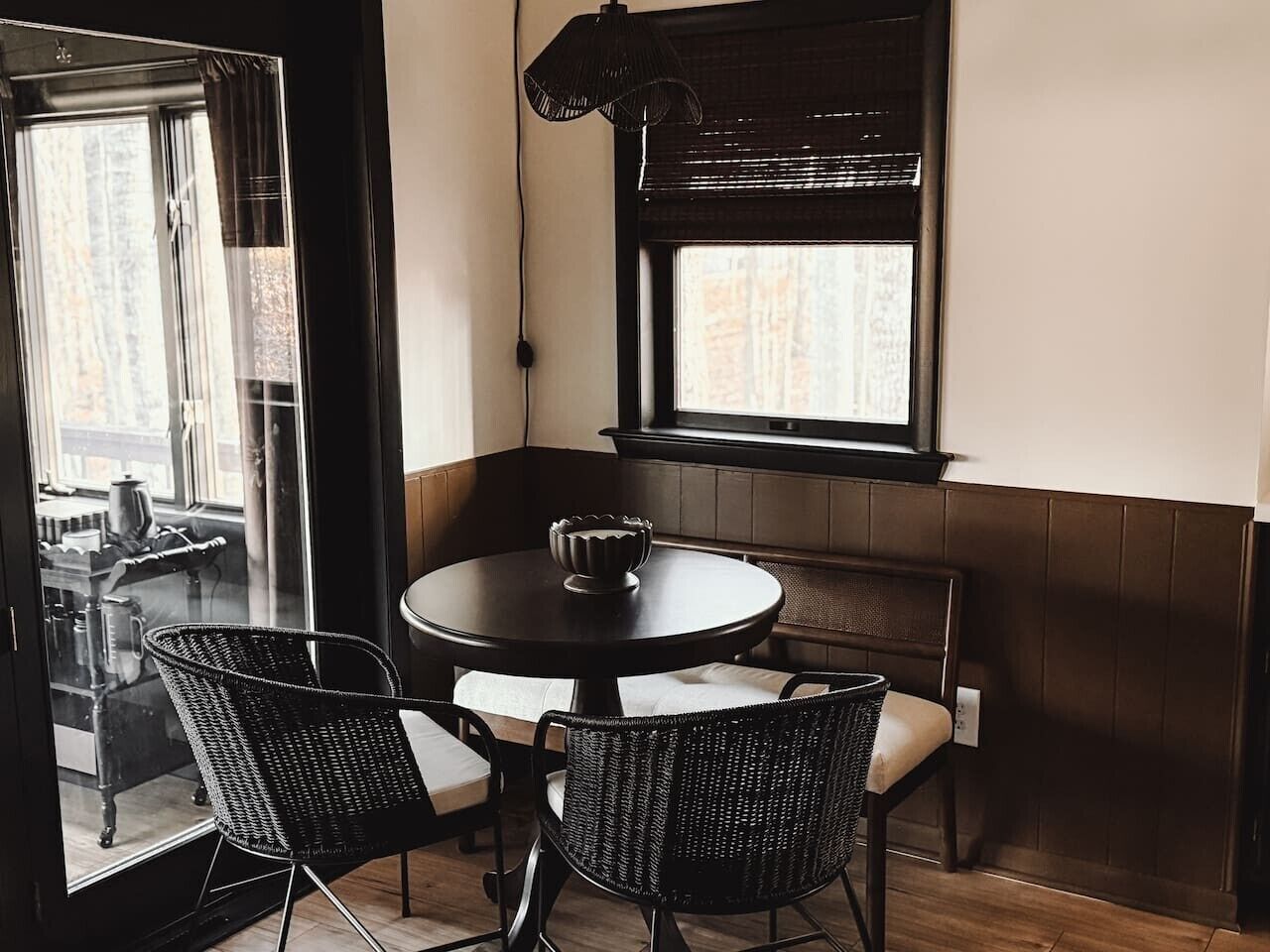 A corner seating area with a round table and two chairs, a window with blinds, and a view of the outside.