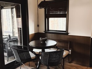 Un coin salon avec une table ronde et deux chaises, une fenêtre avec des stores et une vue sur l’extérieur.
