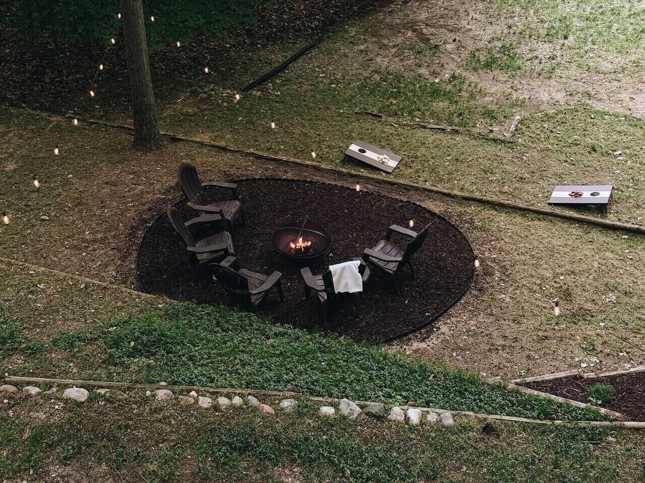 An outdoor seating area with a fire pit surrounded by chairs and a table, set on a grassy lawn with a stone border.