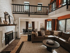A spacious living room with a fireplace, a high ceiling, and a dining area.