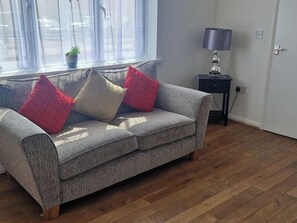 A grey sofa with red and white cushions, a small table with a lamp, and a window with sheer curtains.