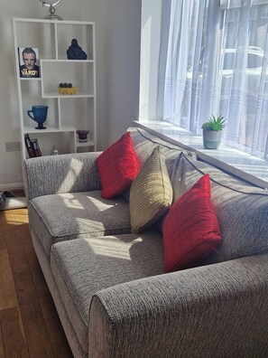 Uma sala de estar com um sofá cinza, almofadas vermelhas e bege, um móvel branco com diversos objetos, e uma janela com cortinas leves.