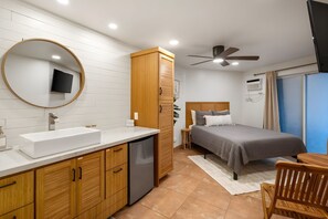A bedroom with a bed, a bedside table, a chair, a ceiling fan, and a bathroom with a sink and mirror. - Kihei, Maui Sunset, Spacious 4 BR, Beachfront, Sleeps 12 (Kihei)