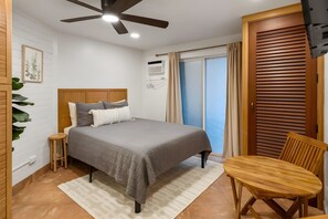 Un dormitorio con una cama, ventilador de techo, cabecera de madera, mesita de noche, una silla y ventana con cortinas.