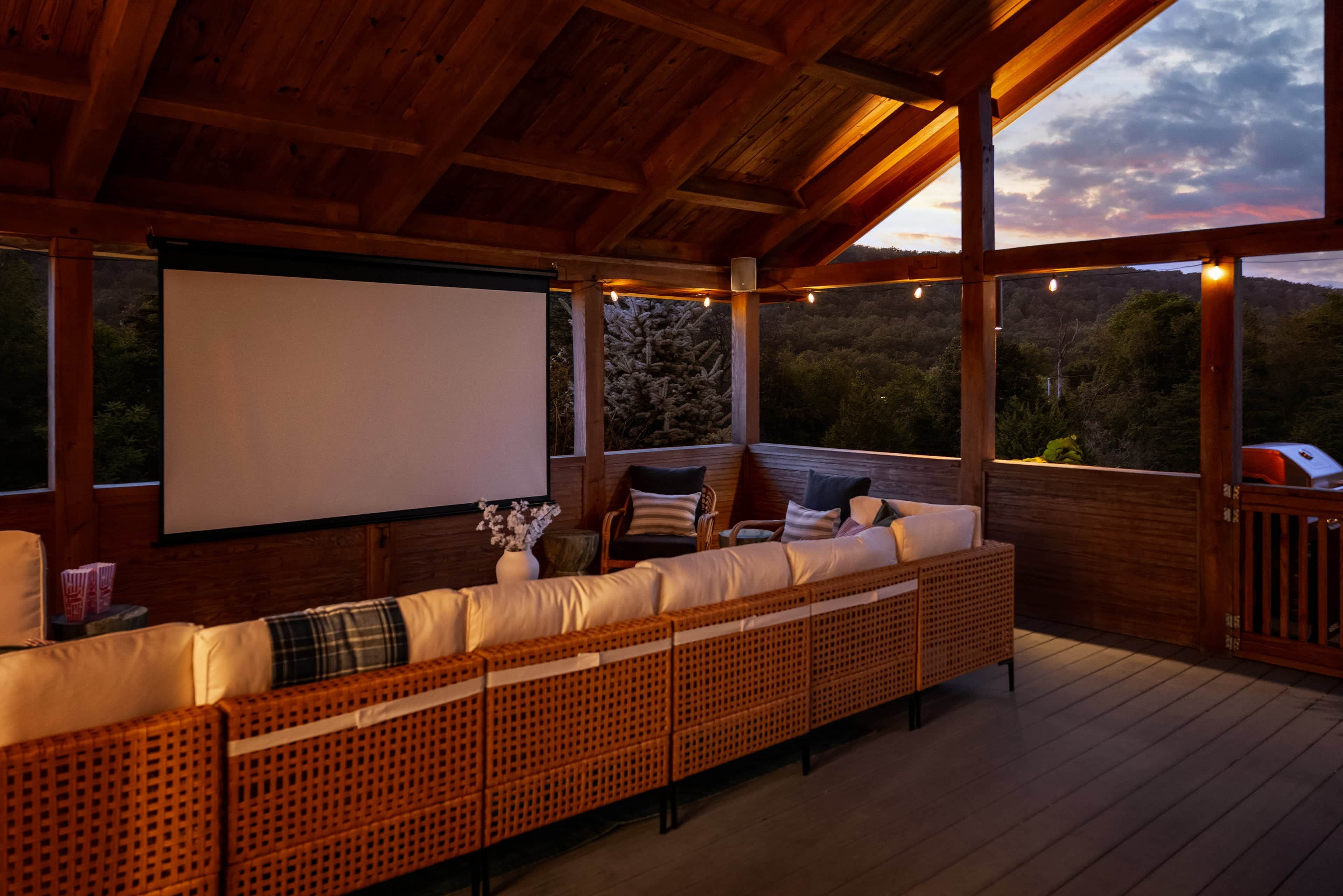 A covered outdoor seating area with a large screen, wooden flooring, and a view of the surrounding landscape.