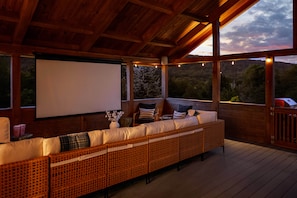 A covered outdoor seating area with a large screen, wooden flooring, and a view of the surrounding landscape. - Stylish & Modern Cabin: Hot Tub, Sauna, Pickleball (Berkeley Springs)