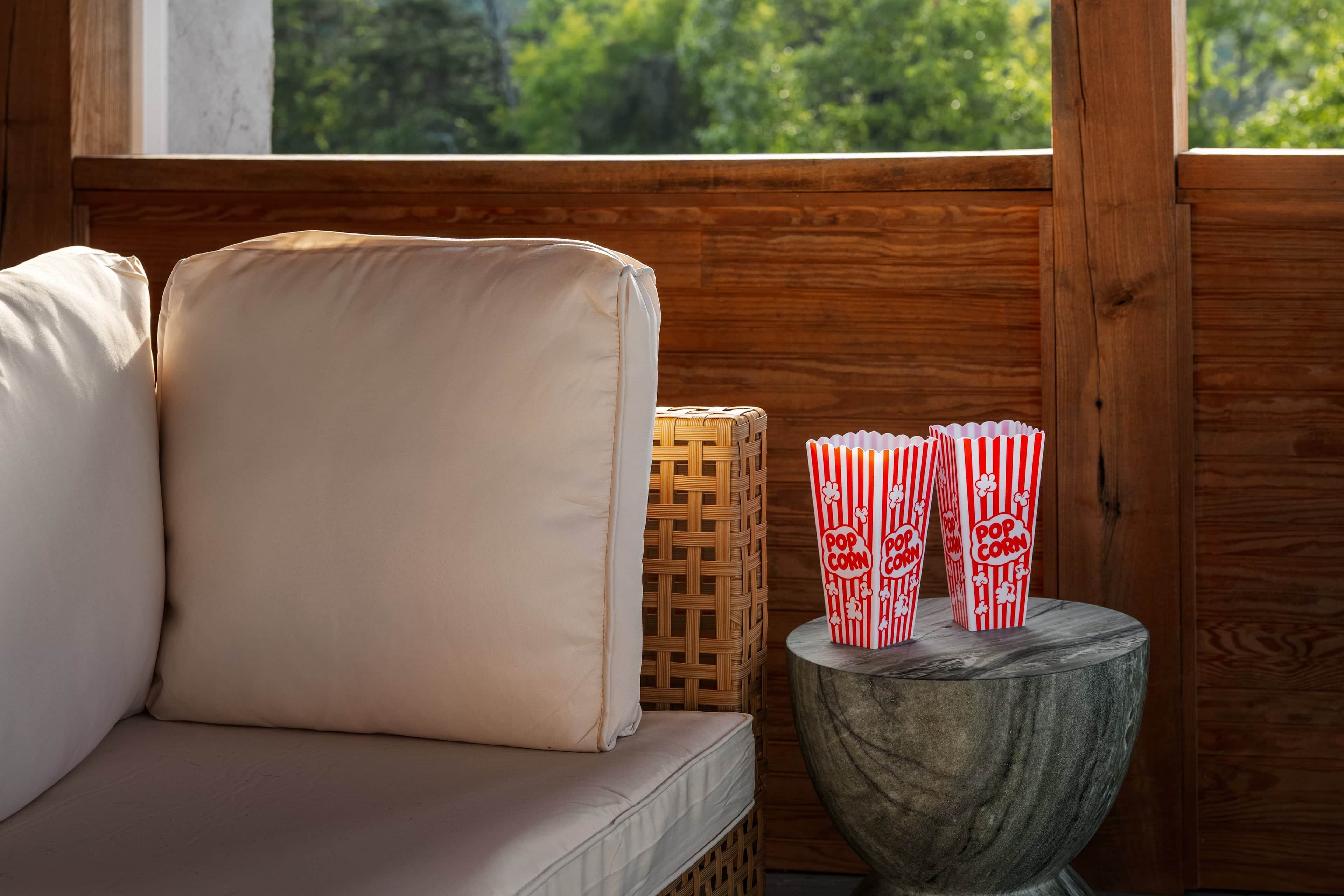 A wooden-framed window with a view of greenery, a cushioned bench, and a small table with striped popcorn containers.