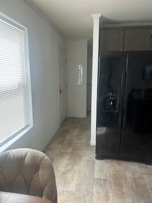 Una cocina con un refrigerador negro, una ventana con persianas y una puerta que da a otra habitación.