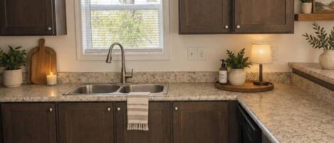 A kitchen with dark wood cabinets, a granite countertop, a window with blinds, a sink, and a small table with a lamp and plants.
