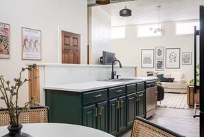 A modern kitchen with dark green cabinets, a white countertop, and a black faucet. There are framed artworks on the walls, a wooden door, and a dining area with wicker chairs. - C-Street Art Loft (Springfield)