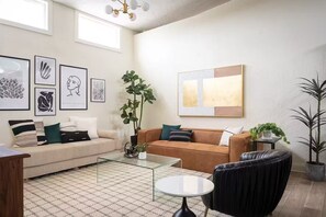 A living room with two sofas, a coffee table, and a black armchair. There are framed artworks on the wall and a large window providing natural light. - C-Street Art Loft (Springfield)