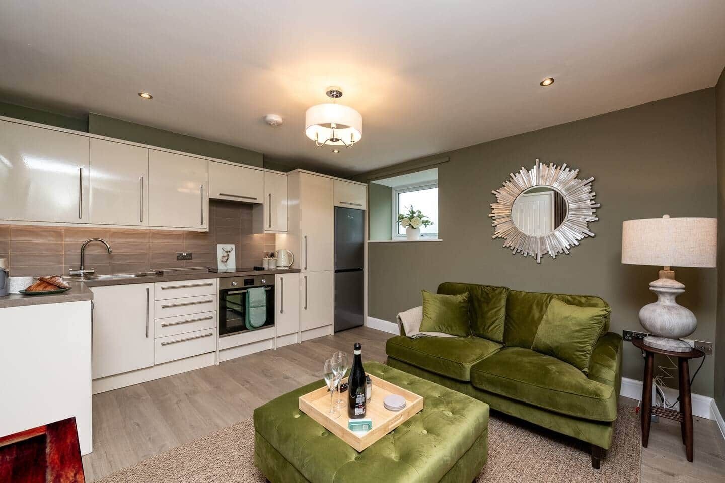 Una cocina moderna con armarios blancos, horno integrado y refrigerador. Hay un sofá verde y una mesa de centro de madera con una bandeja de objetos.
