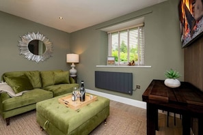 A living room with a green sofa, a wooden coffee table with glasses and a bottle, a lamp, a radiator, and a window with blinds. - No.6 Brantfell House (Bowness-on-Windermere)