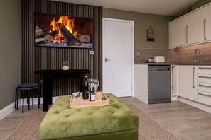 A modern living room with a fireplace, a green ottoman, a dining area, and a kitchen with white cabinets. - No.6 Brantfell House (Bowness-on-Windermere)