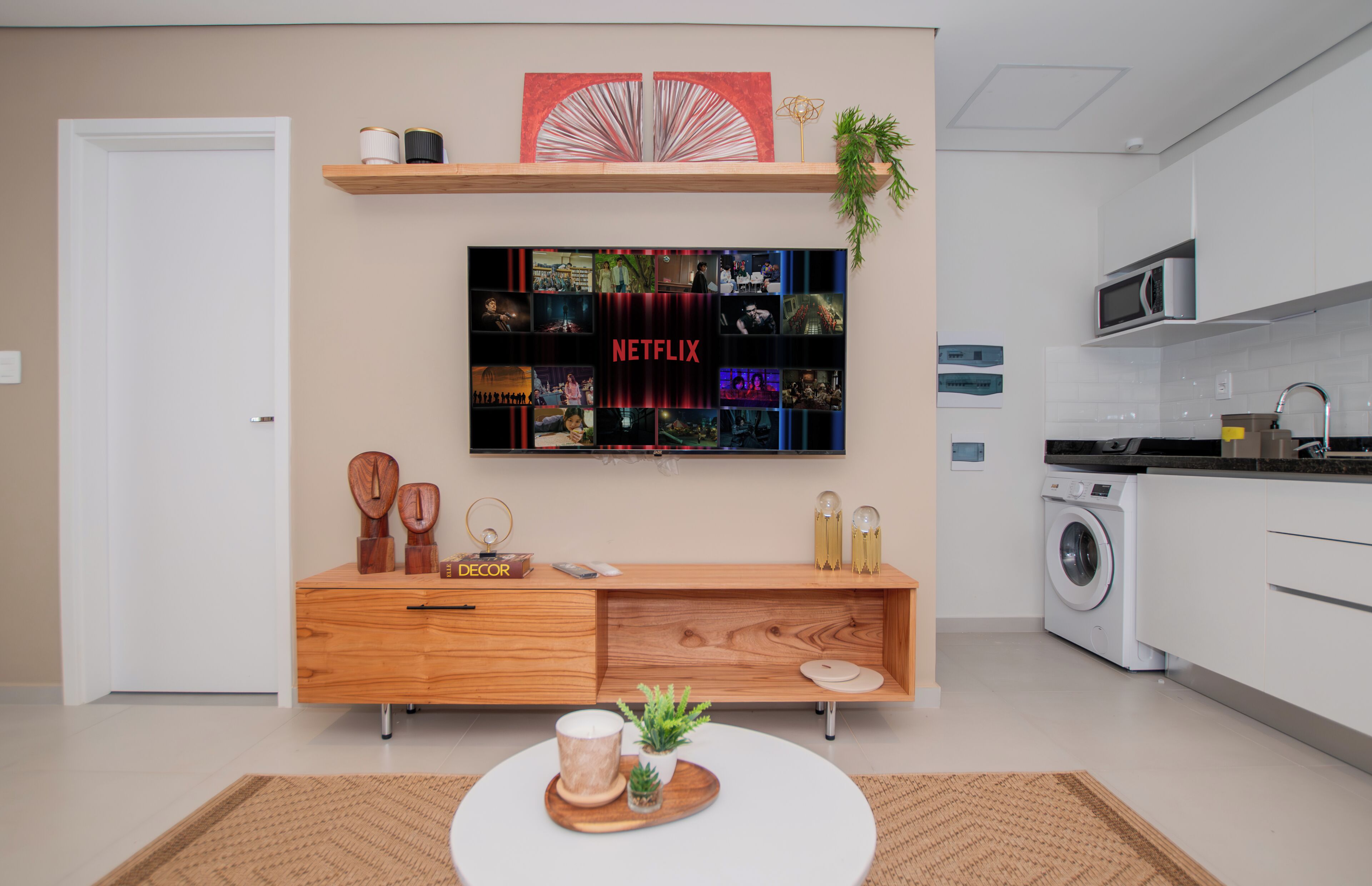 A modern living room with a flat-screen TV mounted on the wall, a wooden console table, a round coffee table with decorative items, and a kitchen area with a washing machine.