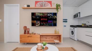 A modern living room with a flat-screen TV mounted on the wall, a wooden console table, a round coffee table with decorative items, and a kitchen area with a washing machine.