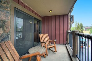 A balcony with two wooden rocking chairs, a glass door, and a view of a building in the background. - It's Four O'Clock Somewhere (Breckenridge)
