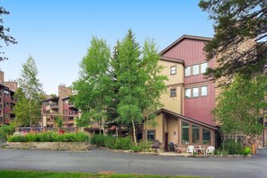 A multi-story residential building with balconies, a parking area, and a small outdoor seating area with chairs and tables. - It's Four O'Clock Somewhere (Breckenridge)