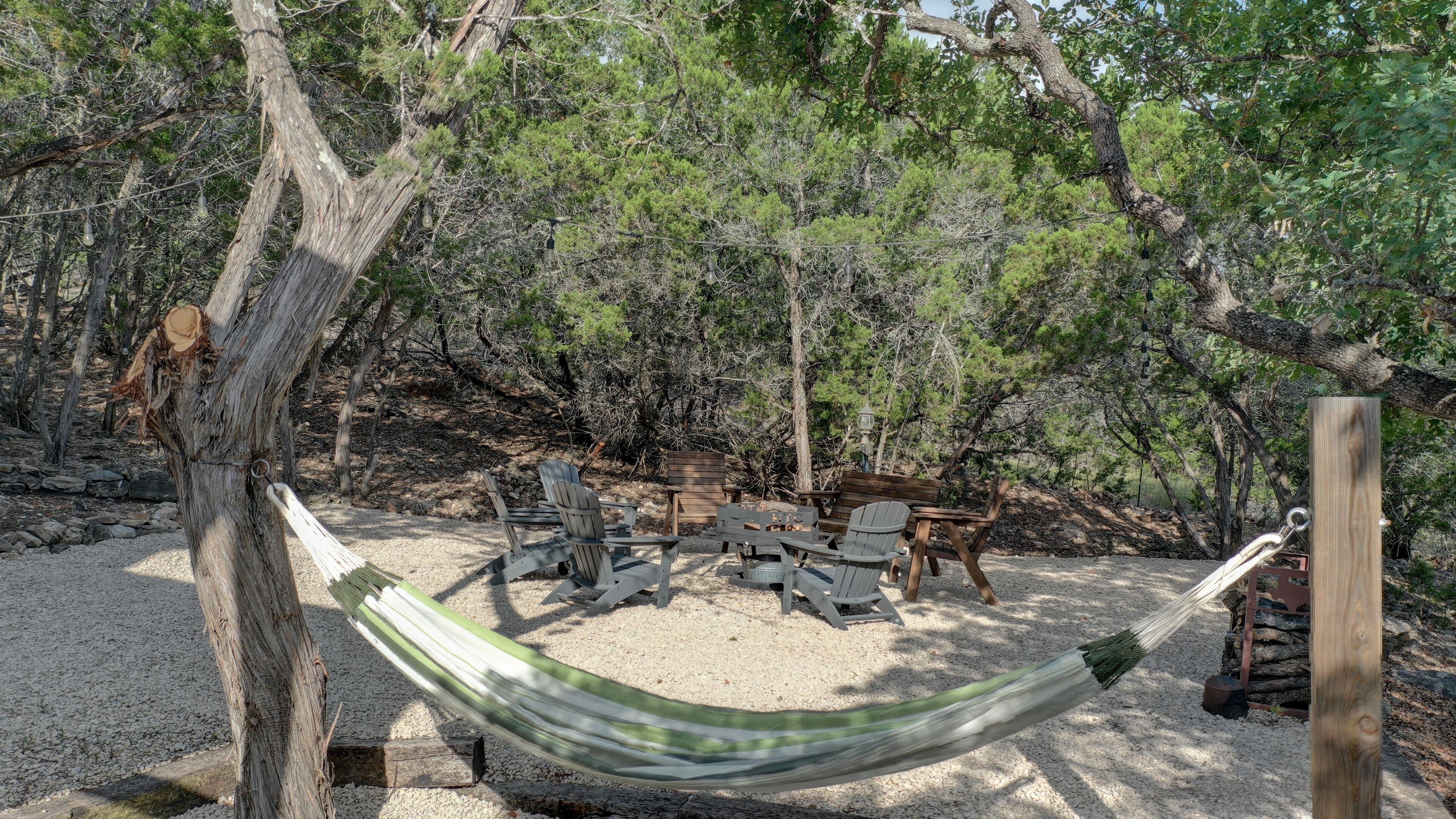 A hammock between trees, surrounded by wooden chairs and a fire pit in a wooded area.