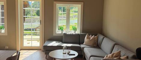 A spacious living room with a high wooden ceiling, a grey sofa, a round coffee table, and two windows with a view of greenery.