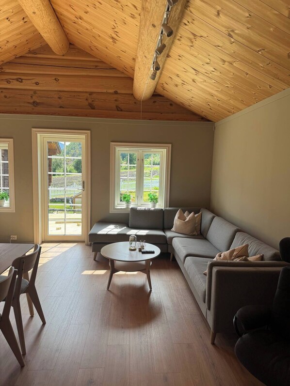 A spacious living room with a high wooden ceiling, a grey sofa, a round coffee table, and two windows with a view of greenery.
