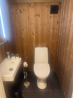A small, wooden-paneled bathroom with a white toilet, a sink, and a toilet paper holder.