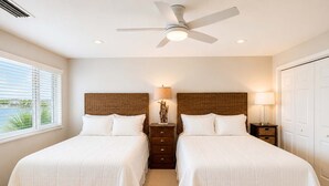 Quarto de hotel com duas camas, ventoinha no teto, mesa de cabeceira com abajur e vista para o exterior.