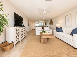 A modern living room with a flat-screen TV, white sofas, a wooden coffee table, and a ceiling fan. - Needle Rush Point E34, Beach Access, 2 Pools, Riverfront, Private Beach (Pensacola)