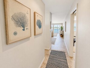 Ein Flur mit zwei gerahmten Kunstwerken an der Wand, einem gemusterten Teppich auf dem Boden und Blick auf einen Balkon mit einer Pflanze.