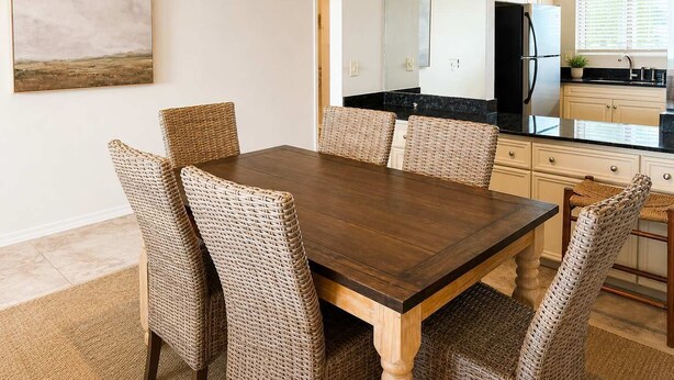 Uma sala de jantar com mesa de madeira e cadeiras de vime, uma cozinha com armários brancos e uma geladeira preta, e um quadro na parede.