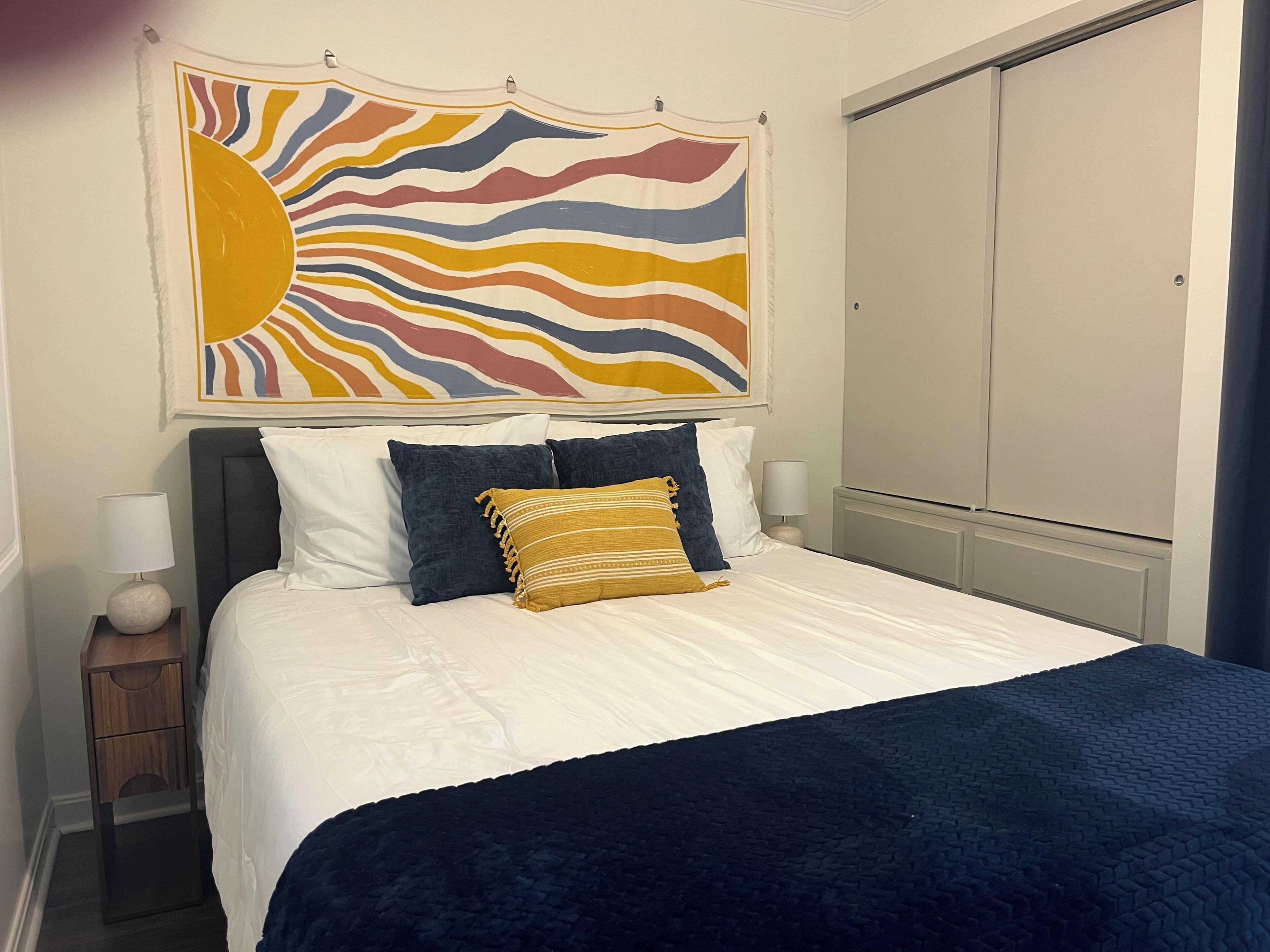 Een hotelkamer met een bed, nachtkastje, lamp, kledingkast en een schilderij aan de muur.