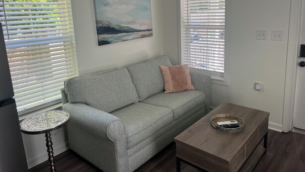 Una sala de estar con un sofá gris, una mesa redonda y una mesa de centro de madera. Hay dos ventanas con persianas, un cuadro en la pared y una puerta con cerradura.