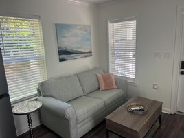 Una sala de estar con un sofá gris, una mesa redonda y una mesa de centro de madera. Hay dos ventanas con persianas, un cuadro en la pared y una puerta con cerradura.