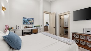 Une chambre moderne avec un grand lit, un bureau, une télévision et une salle de bain visible à travers une porte ouverte.