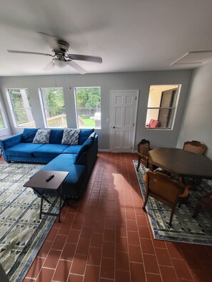 A living room with a blue sofa, a dining table with chairs, a ceiling fan, and a view of the backyard through large windows.